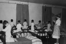 A group of boys in pyjamas kneel on single beds, heads bowed and hands clasped as if in prayer. A woman stands in the room, her hands clasped in the same way.
