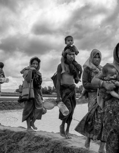 A group of Rohingya women and men carry young children and belongings as they walk in a line over an earthen dike over a stretch of water. Partially obscured.