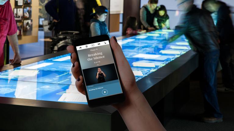 A hand holds up a mobile phone. On the phone are the words “Floor 4” and “Breaking the Silence,” and a person speaking using sign language. In the background, Museum visitors are looking at a large rectangular table with a touchscreen. Partially obscured.