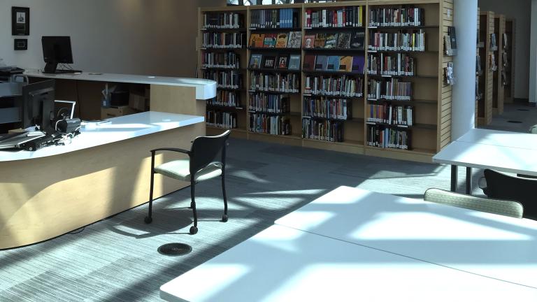 A room containing books on bookshelves, a reference desk with a computer workstation and worktables. Partially obscured.