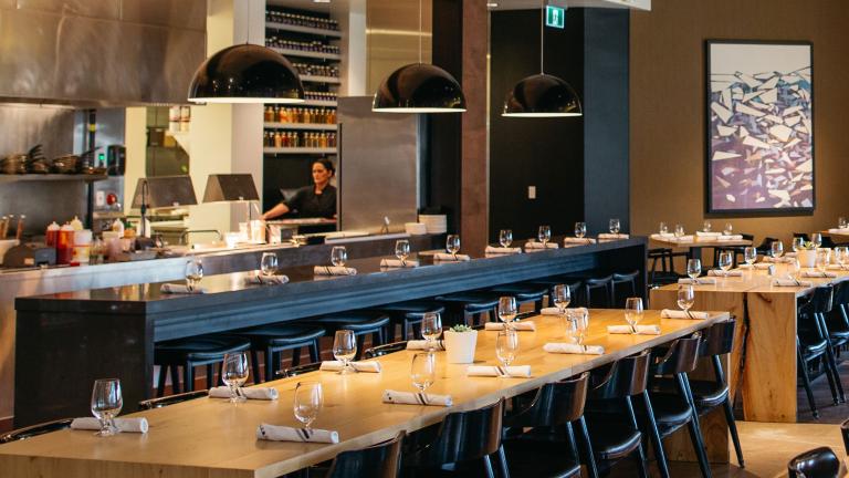 The dining area in a restaurant with long tables and a peek into the kitchen. Partially obscured.