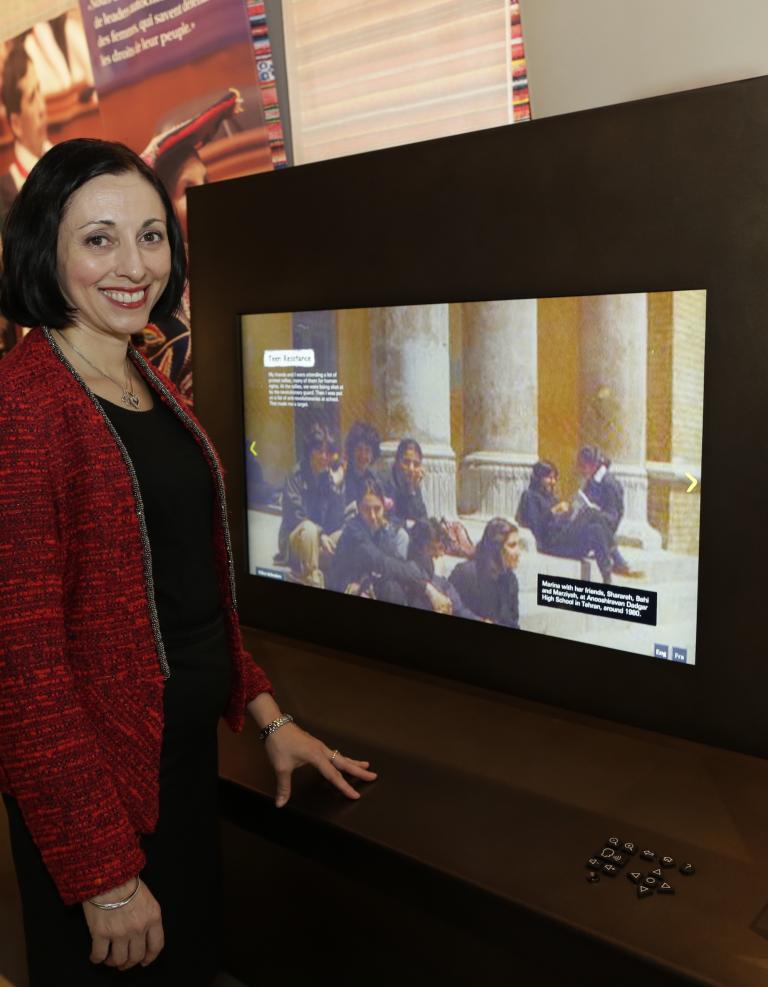 Marina Nemat smiles and stands beside a large digital display screen. She is wearing a red jacket and is looking directly at the photographer. The screen features a picture of people sitting on steps in front of large stone columns. Partially obscured.