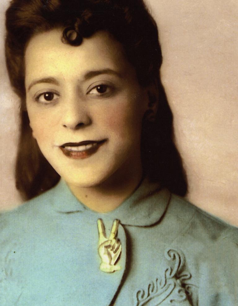 A head-and-shoulder portrait of a smiling Viola Desmond. She is wearing a light blue jacket with embroidered patterns on it. The jacket is held together at the collar by a large pin in the shape of a hand making a “V for victory” symbol. Partially obscured.