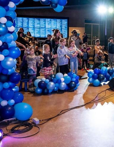 Kids dancing in a hall filled with blue balloons. Partially obscured.
