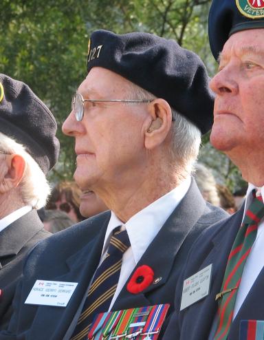 Three older men in Canadian Legion uniforms sit beside each other. They are all wearing Remembrance Day poppies on their uniforms. Partially obscured.
