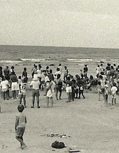 A black-and-white photograph of a crowd of people, most of them standing, on a beach. Partially obscured.