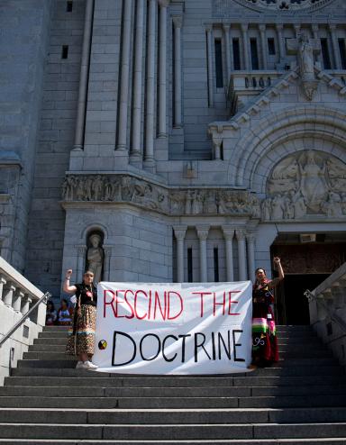 Two people in braids and ribbon skirts raise fists and hold a large cloth banner reading “RESCIND THE DOCTRINE” on the steps of an enormous cathedral. Partially obscured.