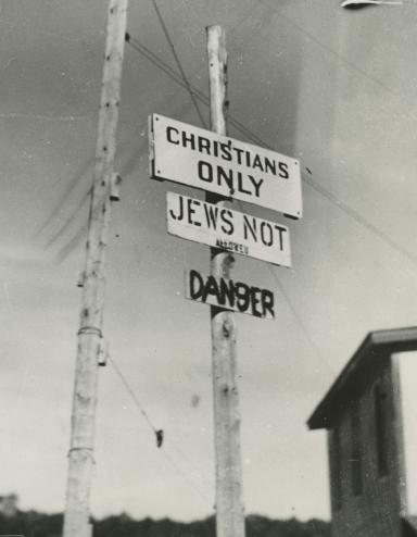 Black and white photo of three signs on a post, reading “Christians only,” “Jews not allowed” and “Danger.” Partially obscured.