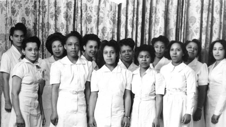 A black and white image of Viola Desmond with 11 other women. All of the women are wearing plain white dresses and are standing in two rows, posing for the camera.