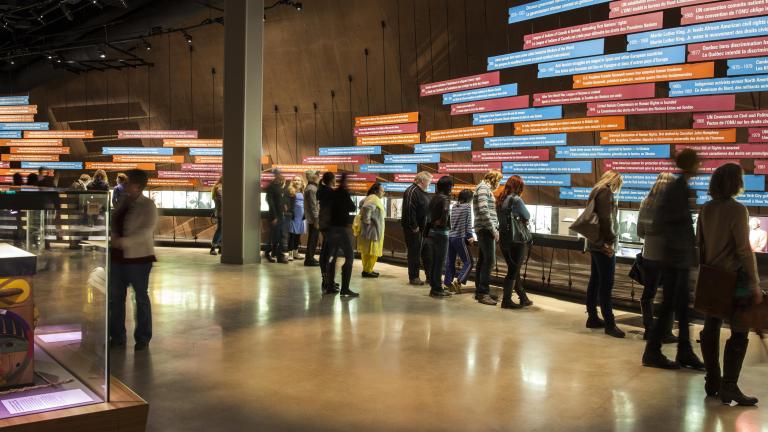 A large crowd in a Museum gallery. Most people are looking at a display on the wall of the gallery. The display consists of a series of pictures and of long rectangular text panels placed horizontally. Partially obscured.
