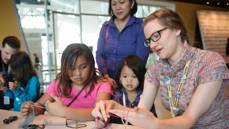 A woman strings yarn around her fingers as she demonstrates and explains what she is doing to two girls. Partially obscured.