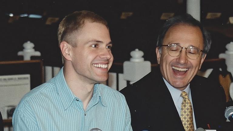 A man wearing a light blue shirt sits at a long table in front of a cluster of microphones, smiling. Seated on both sides of him are other people dressed in suits and facing microphones, also smiling. The name placards along the front of the table read, from left to right, “Murray Billet”, “Robinson Koilpillai”, and “Dr George Rodgers”, with too-blurry-to-read affiliations below the names. Another placard, knocked on its side, reads “Delwin Vriend” without an affiliation. Partially obscured.