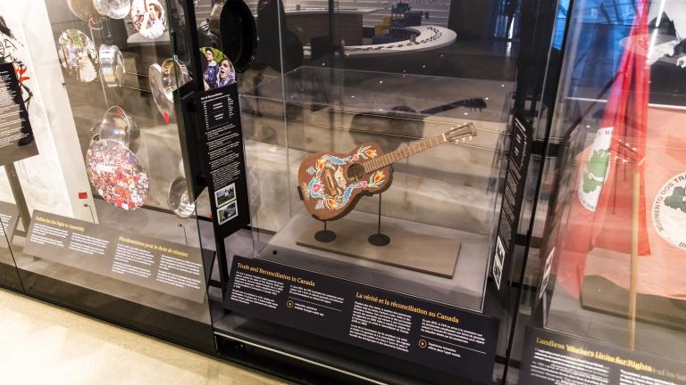 A row of museum display cases with various artifacts inside. A guitar is displayed under glass. The guitar has a colourful floral print with a bird hand-painted onto its front. Partially obscured.